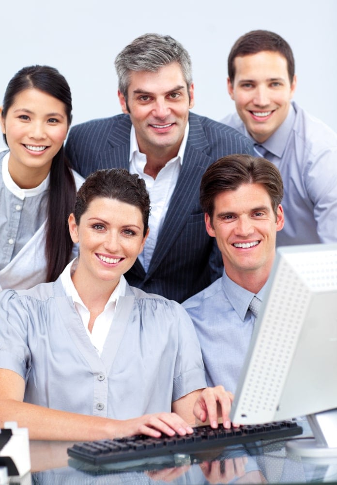 FinCEN real estate report Five professionally dressed people, three standing and two sitting at a desk with a computer monitor, smiling and posing together in an office setting.